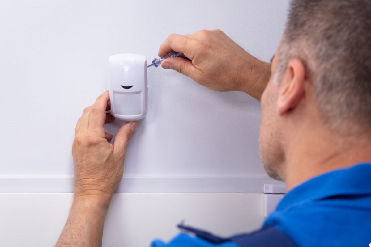 A man adjusts a white alarm mounted on a wall, focusing on the device with tools in hand.